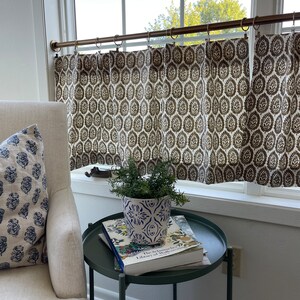 May include: A white armchair with a blue and white patterned pillow sits next to a green side table. The table has a book and a white and blue patterned plant pot with a green plant on top. The window behind the chair has a brown and white patterned curtain with a brass curtain rod.