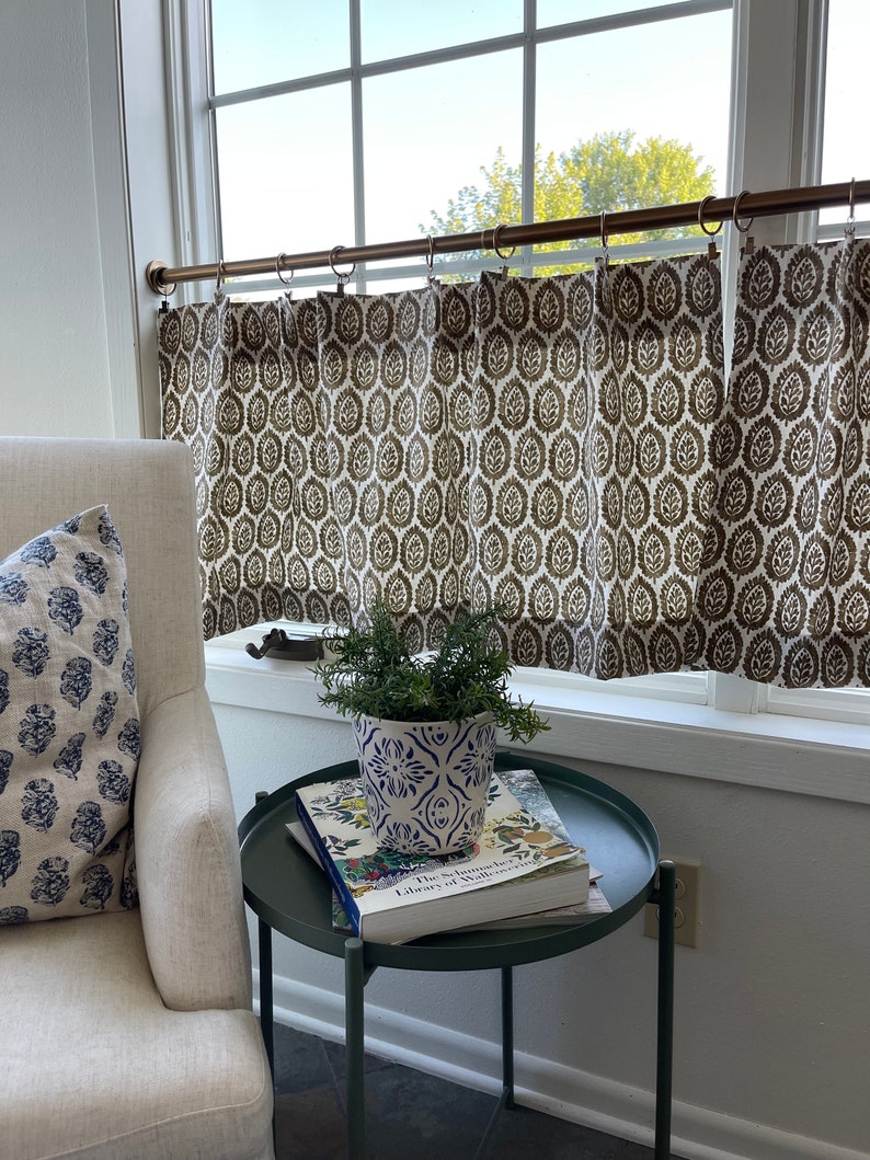 May include: A white armchair with a blue and white patterned pillow sits next to a green side table. The table has a book and a white and blue patterned plant pot with a green plant on top. The window behind the chair has a brown and white patterned curtain with a brass curtain rod.