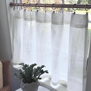 以下が含まれることがあります： 金属リングが付いたブロンズ色のロッドに吊るされた白いリネンのカーテン。カーテンは窓を部分的に覆っており、前景の白いテーブルには小さな鉢植えの植物があります。植物は、灰色の彫刻された頭の形をしたポットに入っています。