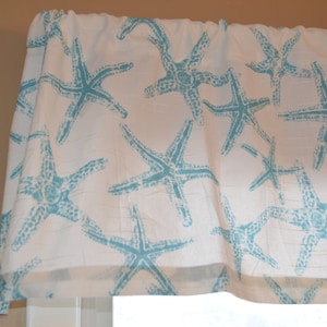 May include: White valance with a repeating pattern of turquoise starfish. The fabric is gathered at the top, creating soft folds. The valance is likely for a window treatment, adding a coastal or beach theme to a room.