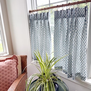Peut inclure: Deux rideaux de fenêtre à motifs blancs et turquoise sont suspendus à une tringle cuivrée. Une plante verte et blanche est posée sur une petite table ronde turquoise devant la fenêtre. Un fauteuil en osier et un coussin rose sont visibles.