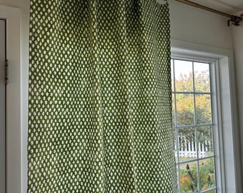 May include: A floor-length curtain featuring a repeating white star-like pattern on a deep green fabric. The curtain is suspended from a metallic bronze-toned rod using circular rings, creating vertical folds across the fabric surface.