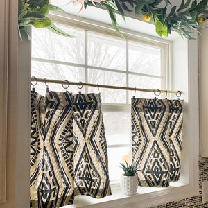 May include: A kitchen window with a gold curtain rod and black and white geometric patterned curtains. The window is decorated with a garland of green leaves and yellow flowers.
