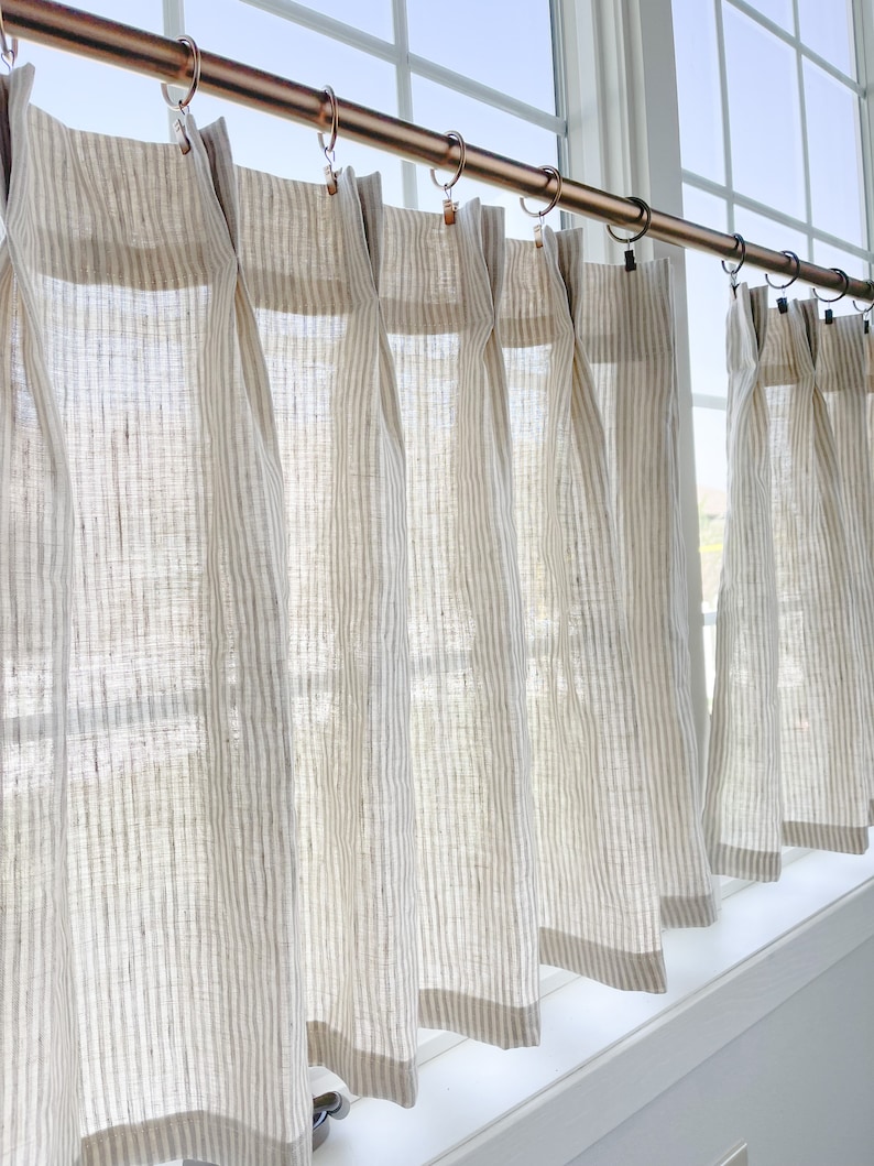 May include: A set of six white linen curtains with thin brown stripes. The curtains are hung on a copper rod with decorative rings. The curtains are gathered at the top and hang straight down to the window sill.