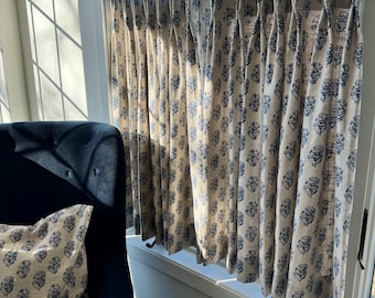 May include: A set of pleated curtains and a matching throw pillow featuring a repeating dark blue floral block print pattern on a light beige fabric. The curtains are hung on a bronze-toned metal rod with circular rings and clips.