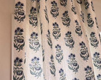 May include: A white curtain panel featuring a repeating floral pattern in shades of dark blue, light blue, and green. The fabric is suspended from a metallic gold curtain rod using circular rings, creating vertical folds across the material.