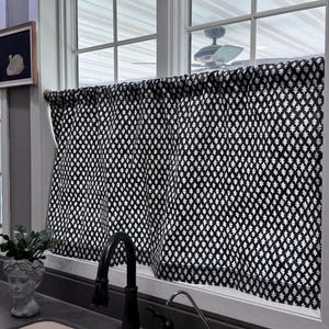 May include: Black and white patterned kitchen curtains hanging in a window. The curtains have a repeating diamond and leaf design. A black faucet and a small decorative plant are visible in the foreground.