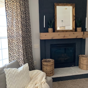 May include: A living room scene featuring a fireplace with a black surround, a wooden mantel, and a gold-framed mirror. Leopard print curtains frame the window. A gray sofa with white pillows and a cream-colored throw blanket with tassels complete the cozy setting.