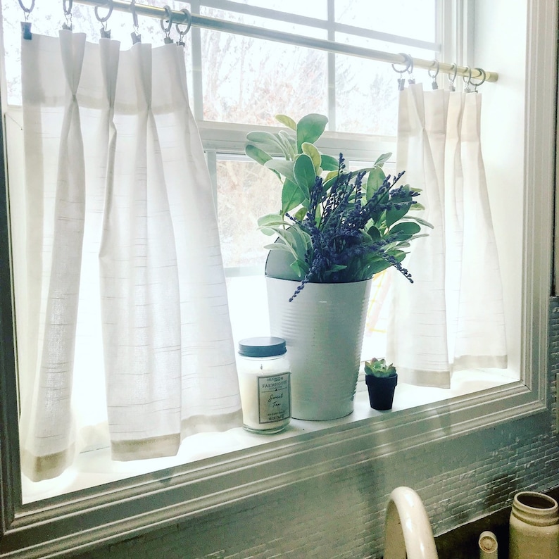 May include: White sheer curtains with a rod hanging above a window. A white metal bucket with a bouquet of purple flowers and green leaves sits on the windowsill. A white candle with a black lid and a black and white label that reads "Farmhouse Fresh" is next to the bucket. A small green succulent plant in a brown pot is on the windowsill.