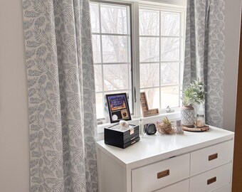 May include: White dresser with six drawers and gold handles. The dresser is in front of a window with white curtains featuring a blue leaf pattern.