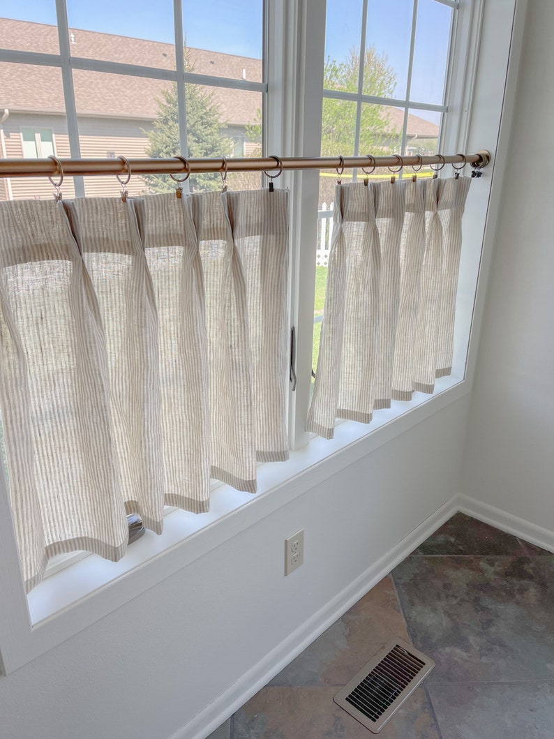 May include: Two sets of white and beige striped curtains with a copper curtain rod. The curtains are hung in a window with a white frame and white trim. The window is in a room with white walls and a tiled floor.