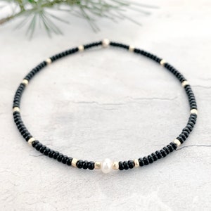 May include: A delicate black beaded bracelet with a single white pearl accent. The bracelet is made with small black beads and gold accents.
