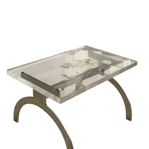 May include: A clear acrylic table with a silver metal base. The table has a rectangular top and the base is made of two curved metal pieces.
