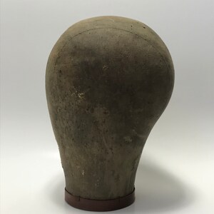 May include: A vintage, brown, fabric-covered, mannequin head with a brown base. The head is slightly tilted to the side.