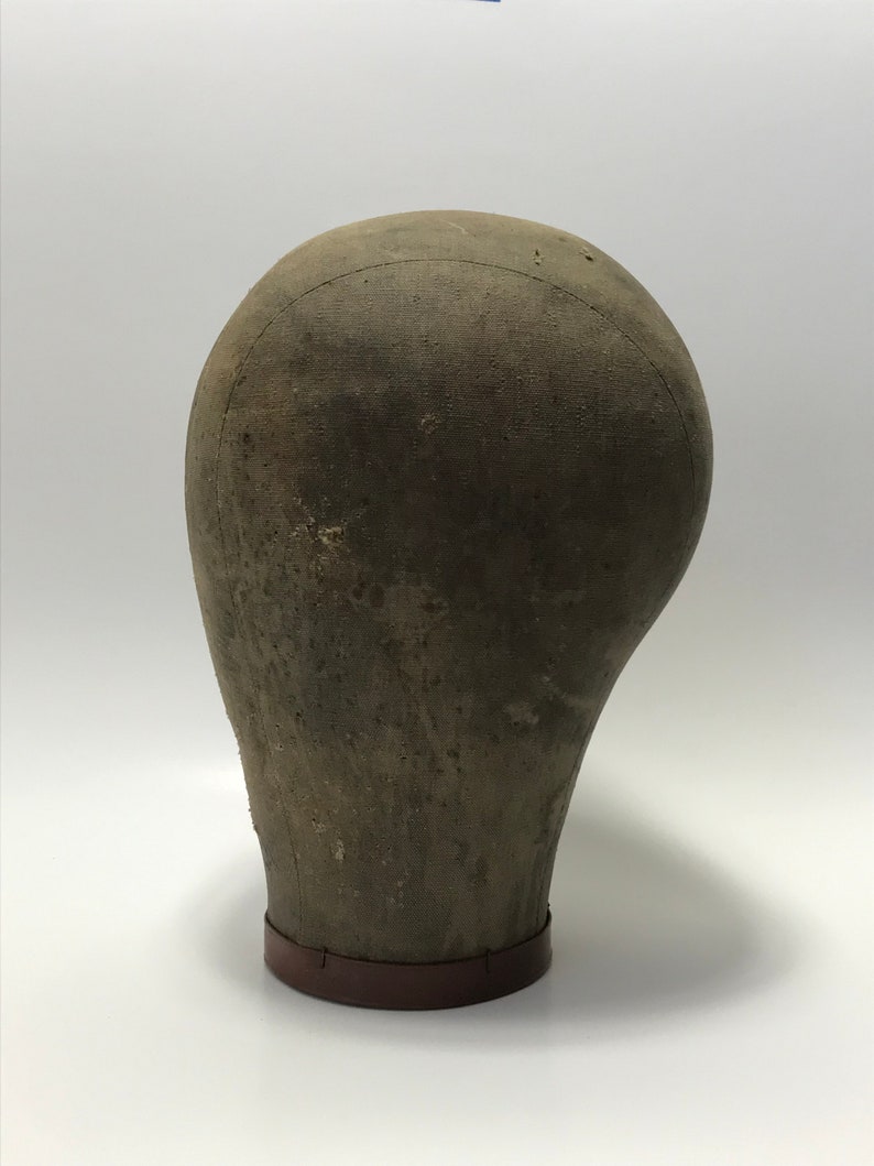 May include: A vintage, brown, fabric-covered, mannequin head with a brown base. The head is slightly tilted to the side.