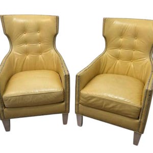 May include: Two yellow leather armchairs with high backs and button tufting. The chairs have a wingback design, nailhead trim, and light-colored legs. The seats are square and the armrests are gently curved.