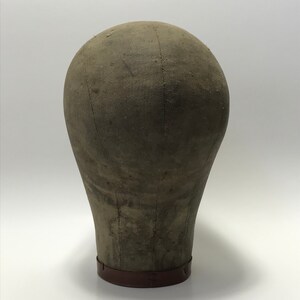 May include: A vintage fabric-covered dressmaker's head form with a brown base. The form is made of a sturdy material and has a slightly rounded shape.