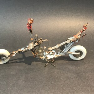 May include: A silver and white metal sculpture of a motorcycle with red wire accents. The motorcycle has two wheels and a long, thin frame.