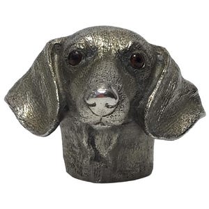 May include: A silver dachshund dog head figurine with brown glass eyes. The figurine is detailed with textured fur and a silver nose.