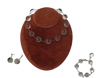 May include: A set of clear glass bead jewelry, including a necklace, bracelet, and earrings. The beads are round and have a silver wire frame. The jewelry is displayed on a brown velvet stand.