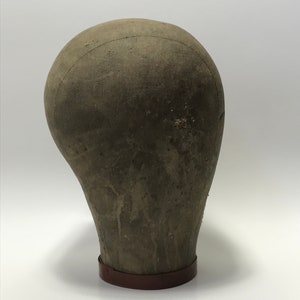 May include: A vintage, brown fabric mannequin head with a brown leather base. The head is covered in a textured fabric and has a worn appearance.