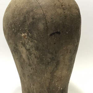 May include: A vintage, brown canvas dressmakers head form with a brown leather base. The form has visible stitching and markings.