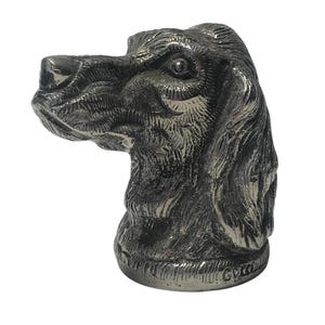 May include: A metallic silver-toned dog head figurine featuring detailed fur texture, long drooping ears, and a prominent snout. The sculpture is cast in a solid material with a polished finish, showing realistic facial features and a circular base.