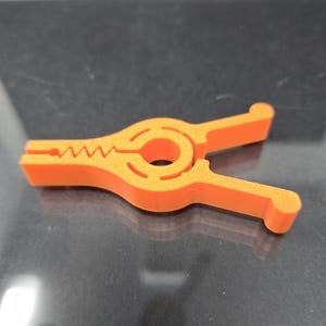 May include: An orange plastic clamp with a circular opening in the center and a toothed gripping surface. The clamp has two handles and is designed to open and close with a squeezing action. The object is likely used for holding items together.