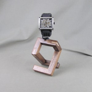 May include: A wooden watch stand with a square design and a light brown wood inlay. A silver watch with a black band is displayed on the stand.