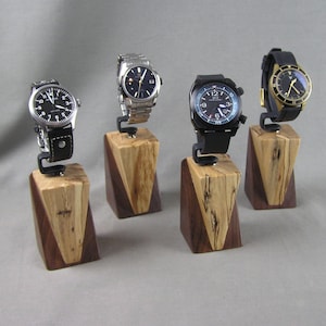 May include: Four wristwatches displayed on wooden stands. The stands are made of light and dark wood with a triangular shape. The watches have black faces and different styles of bands.