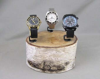May include: Three wristwatches displayed on a wooden watch stand made from birch wood. The stand is round and has a natural bark finish. The watches are all different styles and colors.