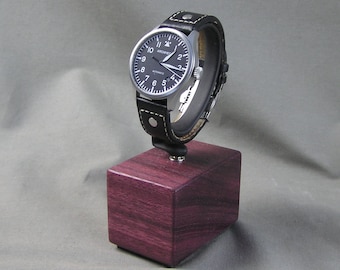 May include: A black-faced wristwatch with white numerals and hands, on a black leather band with white stitching. The watch is displayed on a small, square, purple wooden block. The watch face reads "ARCHIMEDES AUTOMATIC".