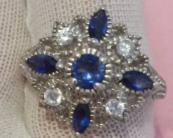 May include: A silver ring with a cluster of blue and white gemstones. The ring features a central blue gemstone surrounded by smaller blue and white gemstones.