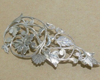 May include: A silver filigree brooch with a floral design. The brooch is made of intricate, delicate silverwork and features a variety of flowers and leaves.