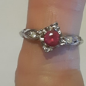 May include: A silver ring with a red gemstone and smaller clear gemstones set in a square design.