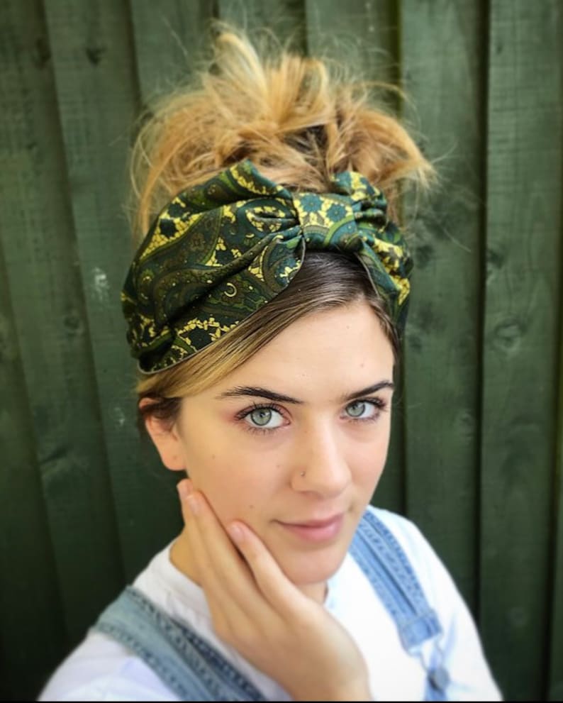 May include: A green and gold paisley patterned headband. The headband is tied in a knot on top of the head. The person is wearing a white shirt and denim overalls.