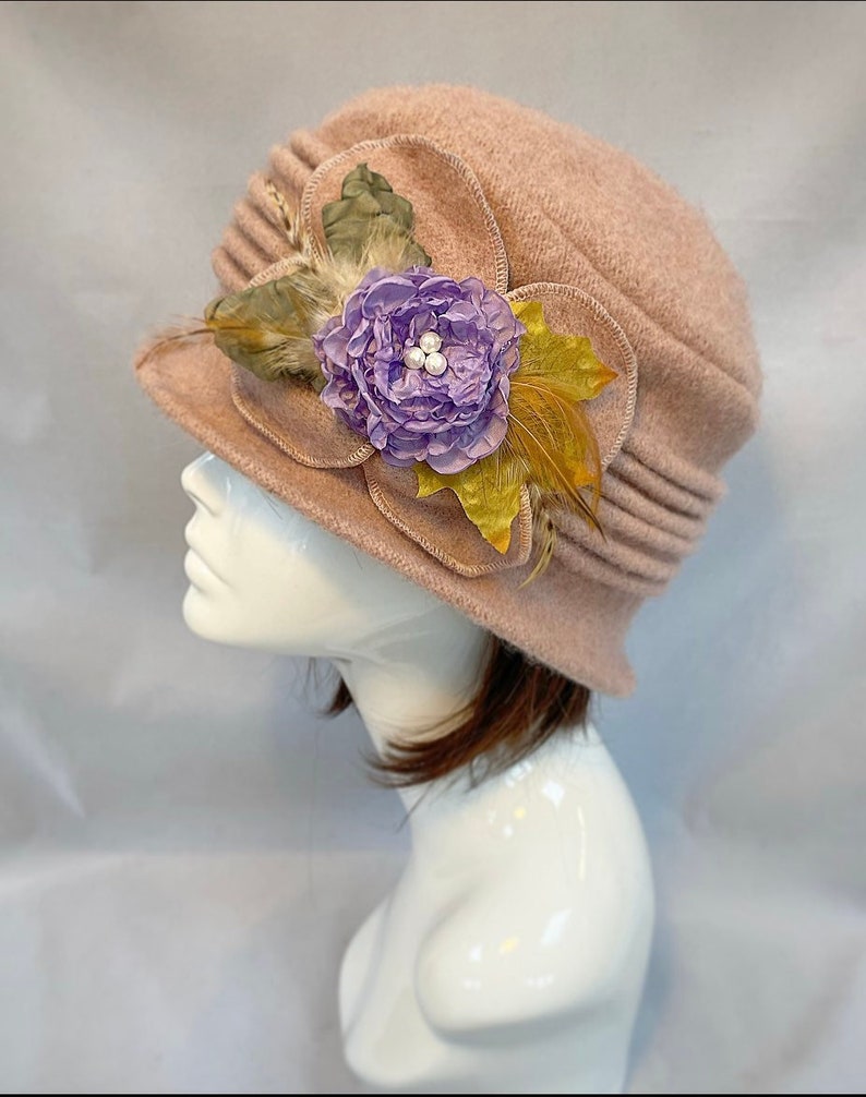 Pu&ograve; includere: Un cappello di feltro rosa con un fiocco di fiori e piume. Il fiore &egrave; viola con accenti di perle bianche e la piuma &egrave; gialla.