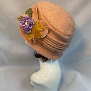 Pu&ograve; includere: Un cappello di feltro rosa chiaro con un fiocco di fiori e piume. Il cappello ha un'ampia tesa e una corona arrotondata.