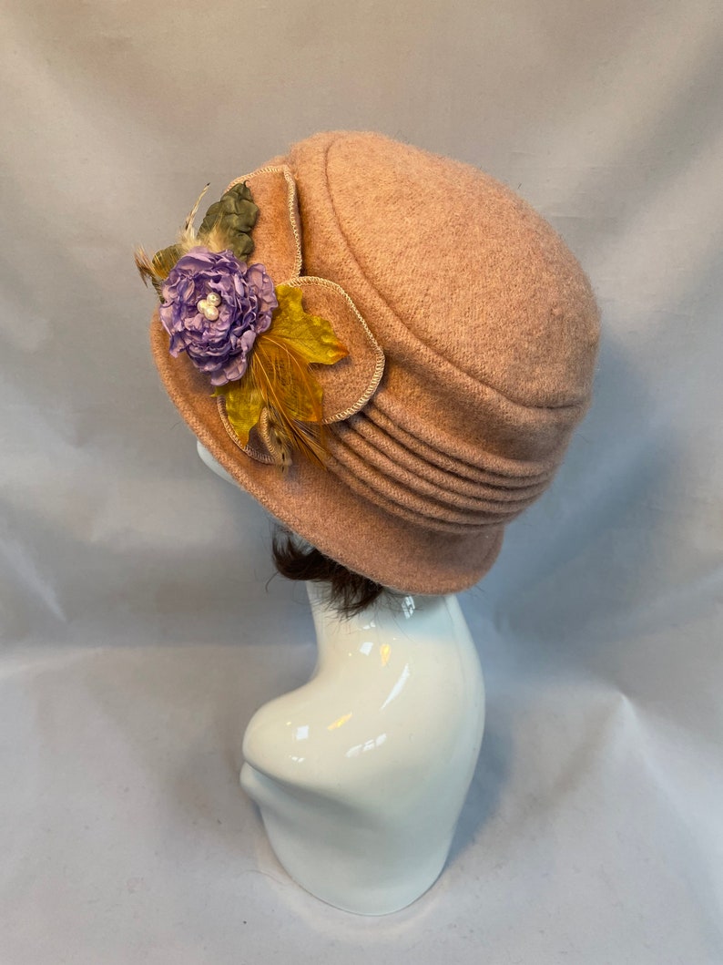 Pu&ograve; includere: Un cappello di feltro rosa chiaro con un fiocco di fiori e piume. Il cappello ha un'ampia tesa e una corona arrotondata.