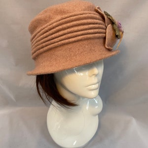 Pu&ograve; includere: Un cappello di feltro rosa con un accento floreale decorativo. Il cappello ha un'ampia tesa e una corona plissettata.