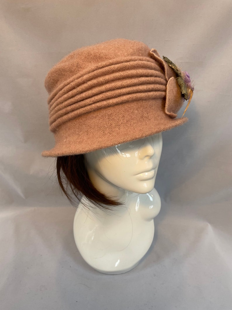 Pu&ograve; includere: Un cappello di feltro rosa con un accento floreale decorativo. Il cappello ha un'ampia tesa e una corona plissettata.