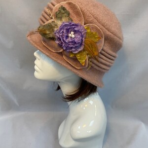 Pu&ograve; includere: Un cappello di feltro rosa con un grande fiore viola e piume sul lato.