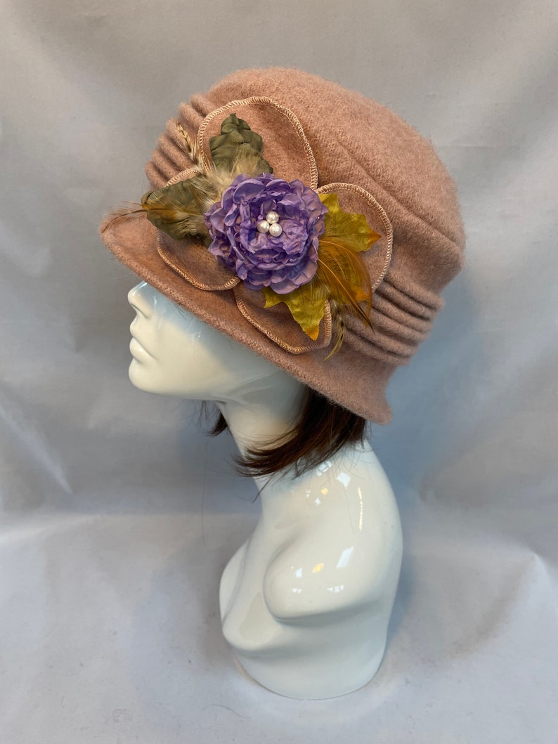 Pu&ograve; includere: Un cappello di feltro rosa con un grande fiore viola e piume sul lato.