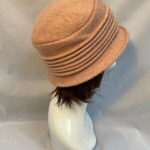Pu&ograve; includere: Un cappello di feltro rosa chiaro con un'ampia tesa e un design plissettato. Il cappello &egrave; su una testa di manichino bianca.