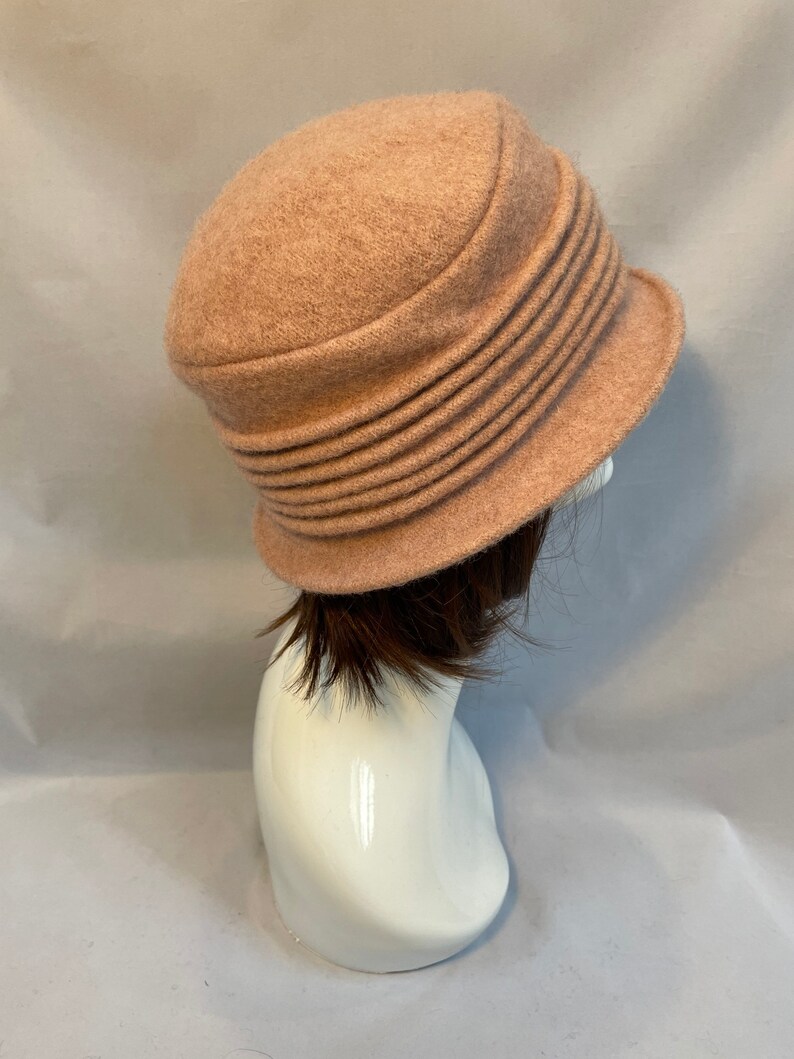 Pu&ograve; includere: Un cappello di feltro rosa chiaro con un'ampia tesa e un design plissettato. Il cappello &egrave; su una testa di manichino bianca.