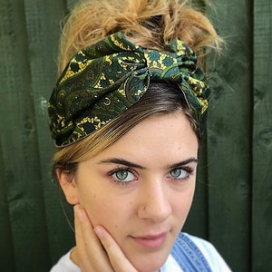 May include: A green and gold paisley patterned headband. The fabric is knotted at the top, creating a stylish accessory. The headband is worn on the head, with blonde hair styled in a messy bun. The background is a dark green wooden fence.