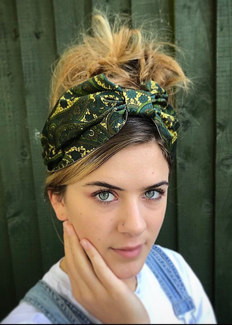 May include: A green and gold paisley patterned headband. The fabric is knotted at the top, creating a stylish accessory. The headband is worn on the head, with blonde hair styled in a messy bun. The background is a dark green wooden fence.