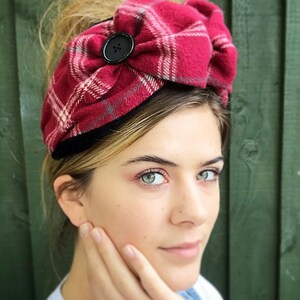 May include: A red and white plaid headband with a black button. The headband is tied in a bow and is worn by a person with blonde hair.