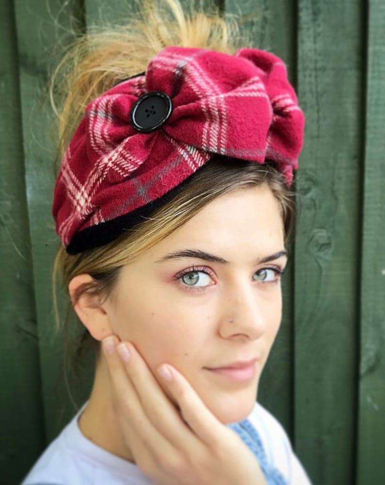 May include: A red and white plaid headband with a black button. The headband is tied in a bow and is worn by a person with blonde hair.
