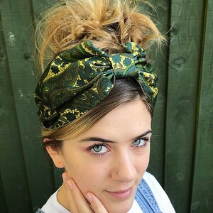 May include: A green and gold paisley patterned headband. The headband is tied in a knot on top of the head. The person in the image is wearing a white t-shirt and denim overalls.